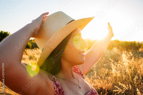 Vászonkép A young woman in a straw hat smiles with her arms raised, basking in warm golden sunset light in a grassy field, evoking summer freedom, joy and peaceful outdoor relaxation
