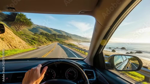 Driver's view along a winding coastal road with hands on the steering wheel, sunlit ocean and cliffs visible through the car interior.