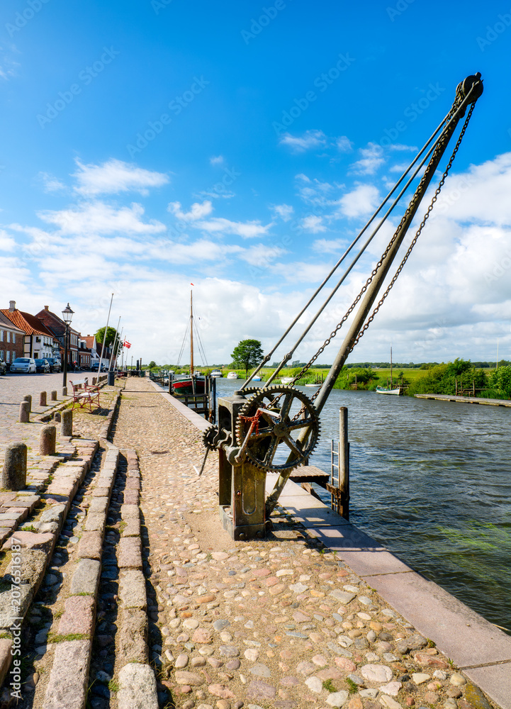 Fototapeta premium Historic harbor crane on Ribe's waterfront with sailing ships, river and traditional harbor buildings beneath a blue sky.