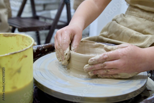 Child hands shaping wet clay on a pottery wheel wearing beige apron in art workshop with yellow bucket, creative educational hobby for skill development and family fun.