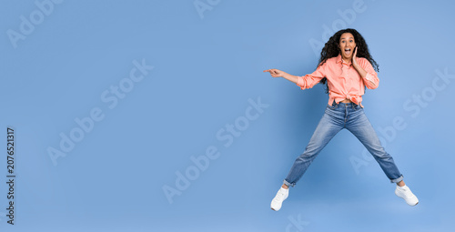 A woman is jumping in the air with a surprised expression on her face. She wears a pink shirt and blue jeans. The background is a solid blue color. This scene takes place during daytime