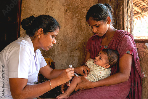 South Indian Female Village Health Worker Vaccinating Infant Held by Young Mother in Rural Hut
