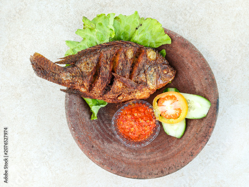 Fried tilapia fish served with red chili, cucumber slices, tomato, and lettuce on traditional clay plate. Indonesian culinary food top view.