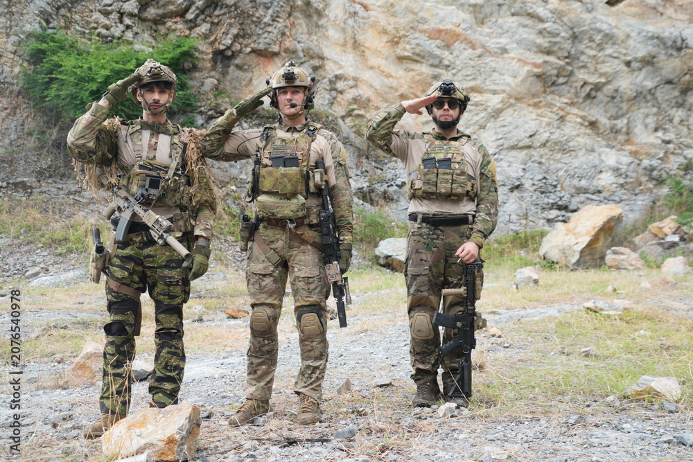 Fototapeta premium Three male soldiers in infantry uniforms are seen holding paintball guns and saluting during a military patrol training exercise in the forest. 