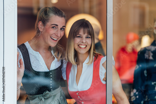 Canvas Print Two smiling women in authentic Bavarian dirndl outfits admire a traditional clothing shop window celebrating local culture and heritage