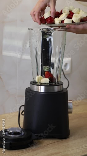 Person putting sliced bananas and fresh strawberries into a glass blender for a healthy drink