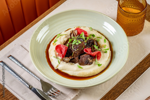 Delicious meal served in a bowl with meat and sauce on mashed potatoes. A dish features meat on top of mashed potatoes with sauce and fresh greens.