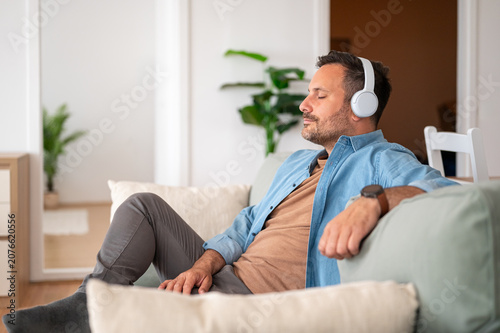 Mature man relaxing at apartment, enjoying sound with wireless headphones, finding peace and comfort by closing eyes and meditating in a tranquil environment