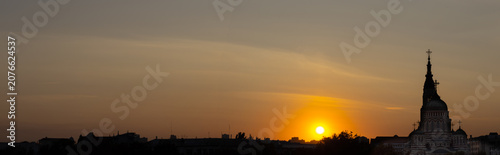The sun sets behind a city skyline. A church tower stands tall against the colorful sky. The scene shows the end of the day with fading light.
