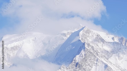 Cinematic close-up video of the snow-covered peaks of the Mont Blanc Massif, with dramatic clouds drifting across the mountains under a clear blue sky. The footage captures the pristine winter