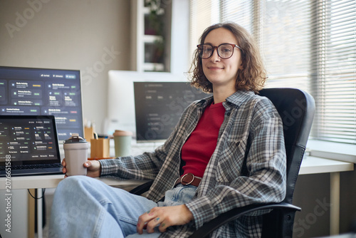 Portrait of young adult Caucasian woman sitting at workstation, holding coffee and monitoring software dashboards on screens. Ideal for tech careers, remote work, productivity
