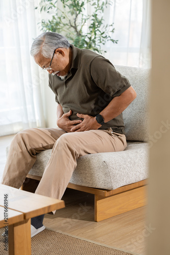 Elderly man stomach pain abdomen sofa living room senior discomfort Elderly man with stomach pain sitting sofa living room, senior male holding abdomen with discomfort near window Vertical