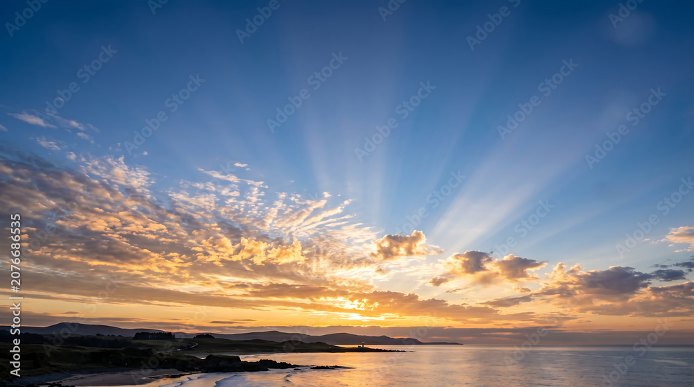 Fototapeta premium Scenic coastal sunset with dramatic sun rays over a tranquil ocean and lighthouse.