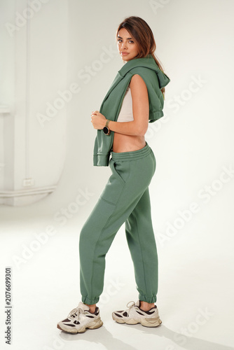 Full-length portrait of a confident young woman wearing a trendy tracksuit and chunky sneakers, standing  in a minimalist studio against a white background.