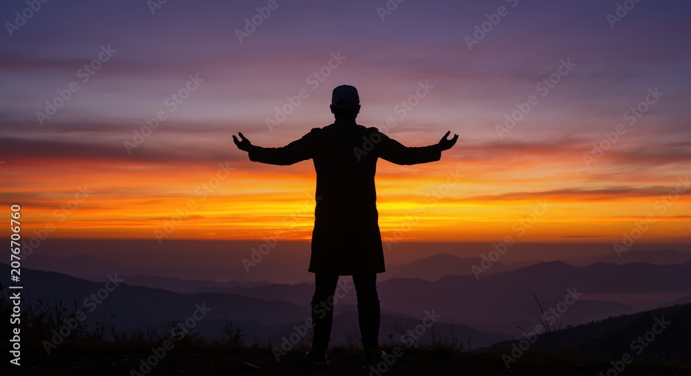 Fototapeta premium Silhouette of Muslim man praying on mountain at sunrise. Person in worship with arms outstretched against colorful sky. Faith and spirituality concept