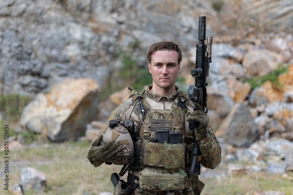 Fototapeta premium Soldier stands in rocky terrain holding a weapon and helmet during a military training