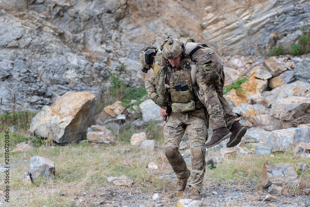 Fototapeta premium Soldier carries injured comrade over rocky terrain
