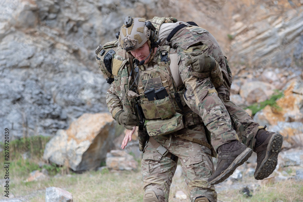 Fototapeta premium Soldier carries injured comrade over rocky terrain