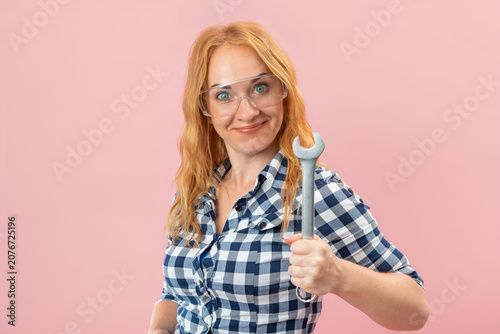Smiling woman wearing safety goggles and a plaid shirt, confidently holding a wrench and looking at the camera against a pink background, representing skill and readiness for work