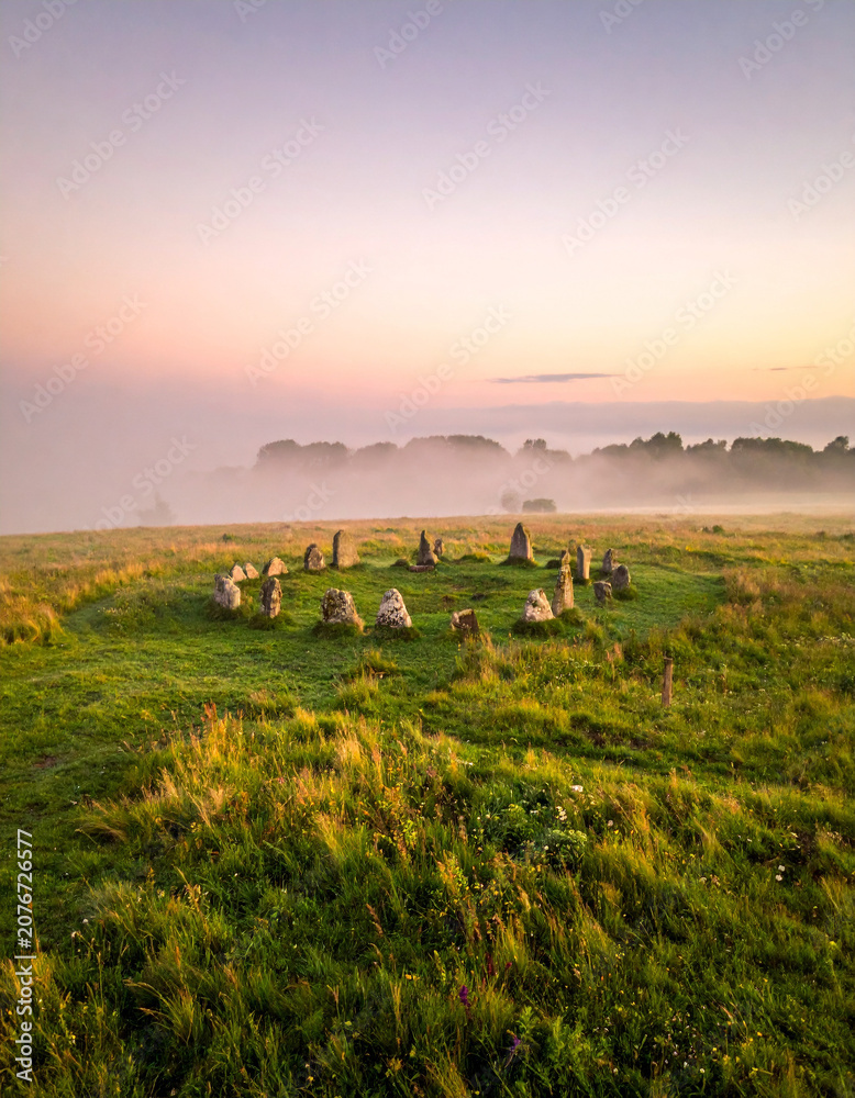 Fototapeta premium Morning mist rolls over a prehistoric stone circle standing in a lush green meadow within the English countryside landscape.
