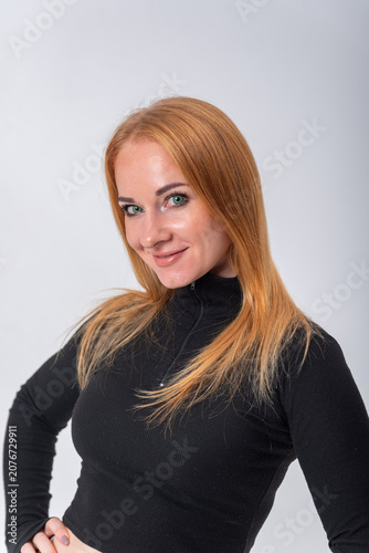 Young woman with red hair and green eyes confidently smiling, looking directly at the camera while posing in a black long sleeve top on a clean studio background