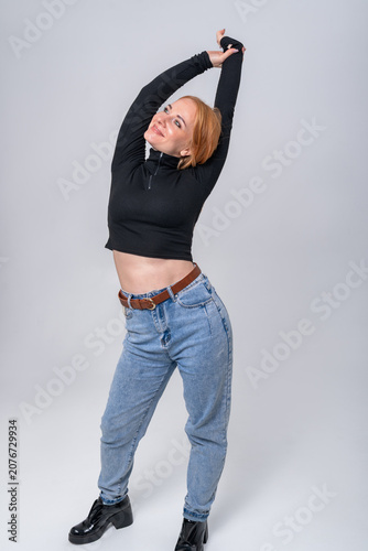 Young woman with red hair stretching her body to the side, expressing happiness and carefree attitude in a casual outfit, standing on a light background