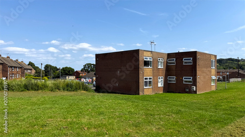 A red brick building rises from a broad grassy field beneath a clear blue sky, with quiet homes and lush greenery surrounding it in Thirsk Grange, Clayton, Bradford, UK