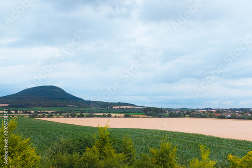 Bohemian Central Highlands, Czech Republic: solitary volcanic hill mountain behind green agricultural crop and golden wheat fields, rural vista.