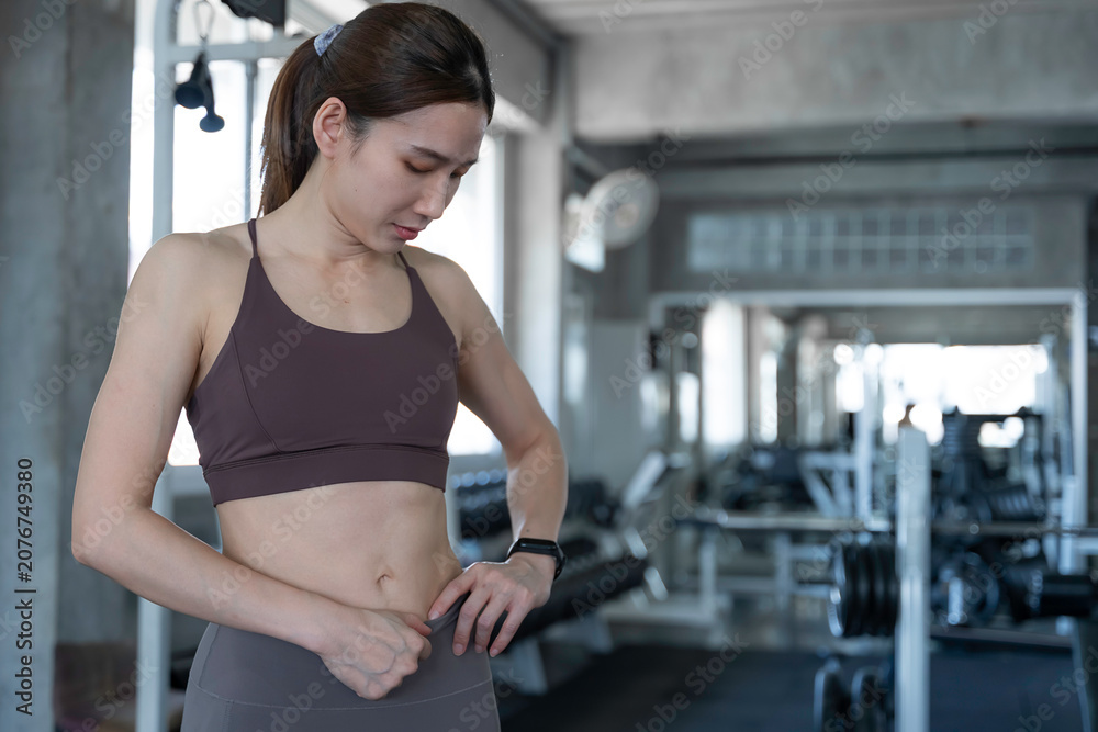Fototapeta premium Happy confident Asian woman showing her toned abs after a workout at the modern fitness center promoting a healthy active lifestyle and physical wellbeing for diverse health campaigns.