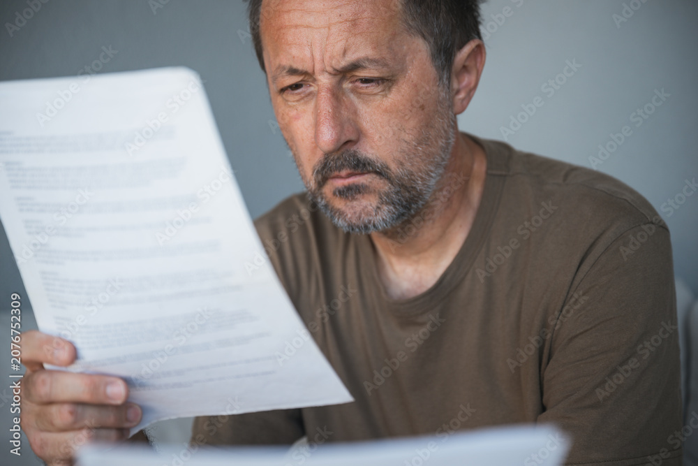 Fototapeta premium Middle-aged man studies a printed document intently, hand on chin and furrowed brow conveying worry, concentration and contemplation while reading important paperwork at home.