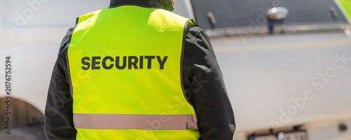 Rear view of security officer wearing yellow reflective vest labeled SECURITY while observing people in sunny urban setting
