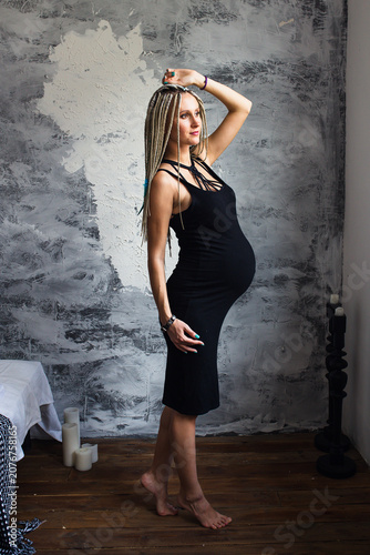 Beautiful pregnant woman with dreadlocks wearing a black dress near a gray wall at home, tender pregnancy lifestyle portrait.