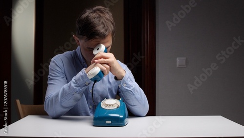 Fotografija Man in blue shirt sitting at a table with a vintage rotary phone, experiencing s