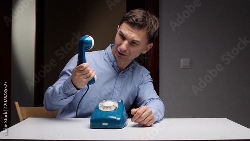 Fotografie Man in a blue shirt sitting at a table, holding a vintage blue rotary phone rece