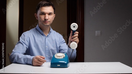 Billede på lærred Young man in a blue shirt holding a retro telephone receiver, looking directly a