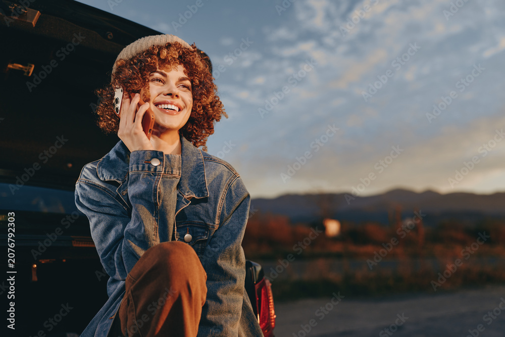 Obraz premium Woman lifestyle moment, smiling woman wearing denim jacket, holding phone to ear, curly hair, warm sunset sky, outdoors in evening air, relaxed casual travel mood, city background blur