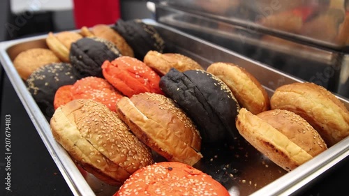 Commercial stainless steel tray featuring assorted black, red, and golden sesame burger buns, placed beside a chafing dish on a black tablecloth for a buffet event and catering service.