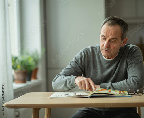 Senior Man Pointing and Reading Children's Book At Wooden Table