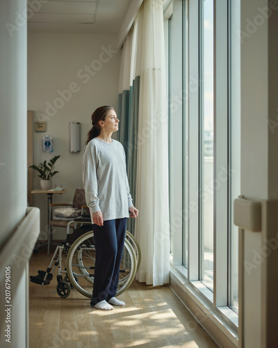 Recovering Female Patient Standing by Window Next to Wheelchair in Hospital
