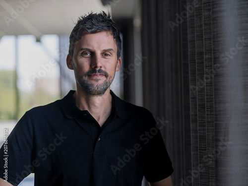 Office portrait of a professional man with a calm, closed-mouth expression beside a daylit window.