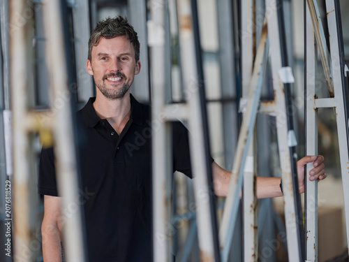 Bilde på lerret Warehouse worker grips a vertical frame and smiles broadly, framed by racks in a warehouse