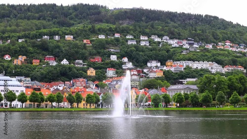 Fountain sprays water in perfect arc across calm lake surface. Colorful houses line waterfront, nestled against green hills. Trees frame picturesque scene with gentle breeze rustling leaves