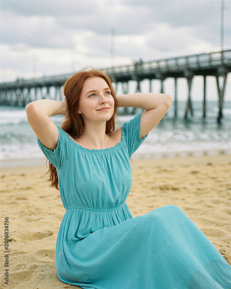 Fototapeta premium A young red-haired woman in a blue dress sitting on a sandy beach, smiling, with a pier stretching into the water behind.