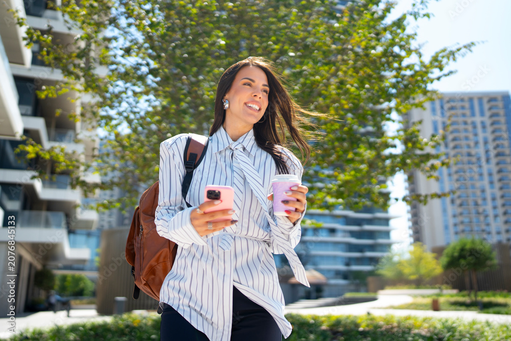 Obraz premium A woman walks outdoors with two coffee cups in her hands. She is smiling and looking at her phone while wearing a backpack. The scene is in a city with tall buildings and greenery.