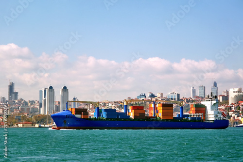 Self-geared container ship sailing past Beşiktaş with Istanbul skyline of high-rise towers and construction cranes in background