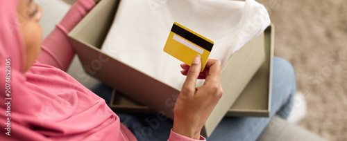 Tableau sur toile Excited Young Asian Woman Unboxing New Clothing Package While Holding Credit Card In Home Setting, Anticipating Shopping Online Experience