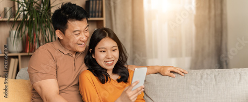Fotografía Happy Asian Couple, Young Adults, Sitting Close Together On Couch, Sharing Laughter While Using Smartphone At Home, Enjoying Time Together In Cozy Living Room