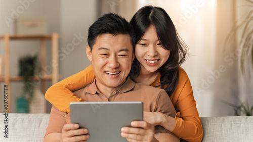 Obraz na płótnie Happy Asian Couple Smiling Together While Holding Tablet And Enjoying Time Together At Home, Sitting On Couch In Cozy Living Room
