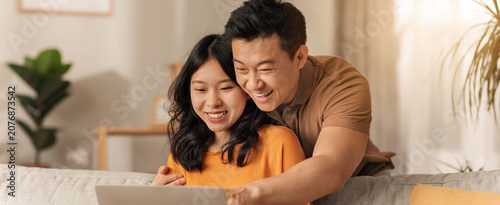 Foto Happy Asian Couple Smiling While Using Laptop Together At Home, Sitting On Couch In Cozy Living Room