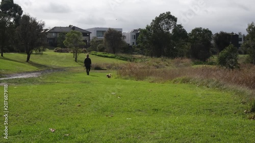Person walking a small dog through green parkland corridor in Caroline Springs, Melbourne, Australia. Suburban open space. Concept of active lifestyle, wellbeing, leisure and community liveability